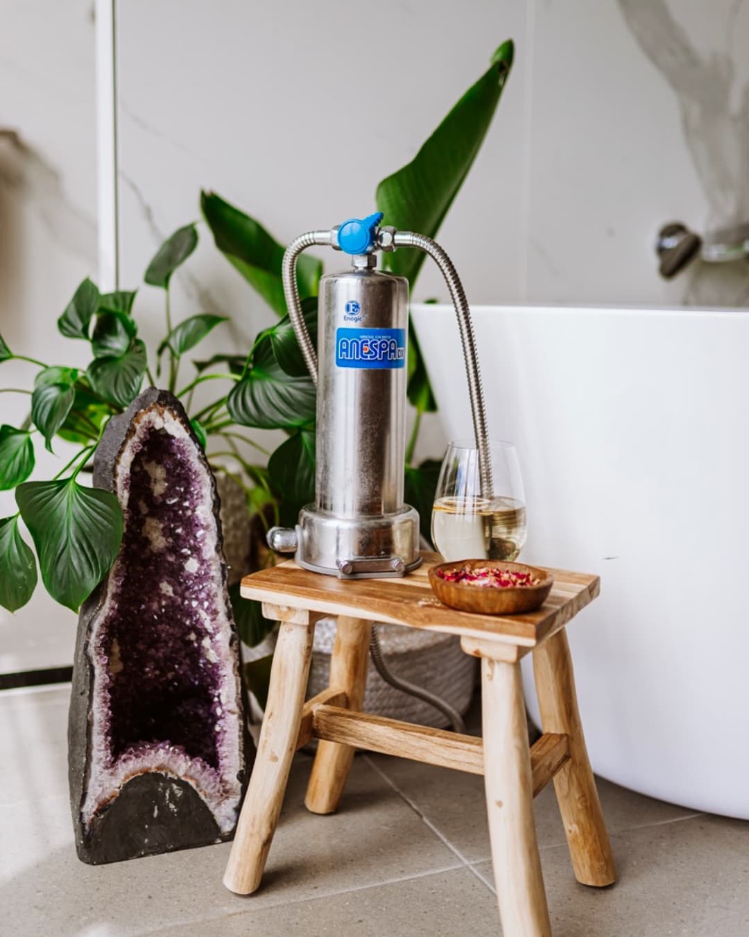 Enagic Anespa DX shower and bath mineralisation unit on a stool beside a freestanding soaking tub, styled with plants and an amethyst crystal