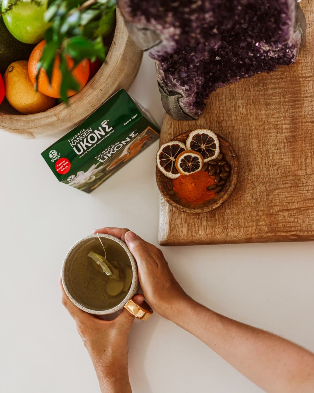 Overhead view of hands holding turmeric tea beside a Kangen Ukon supplement box, dried citrus, and turmeric powder on a wooden board